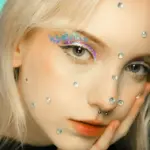 Lähikuva naisen kasvoista Studio Lumon kauneuspalveluissa, jossa kimaltava glitter-luomiväri ja suuret timanttikimalteet koristavat koko kasvojen ihoa.