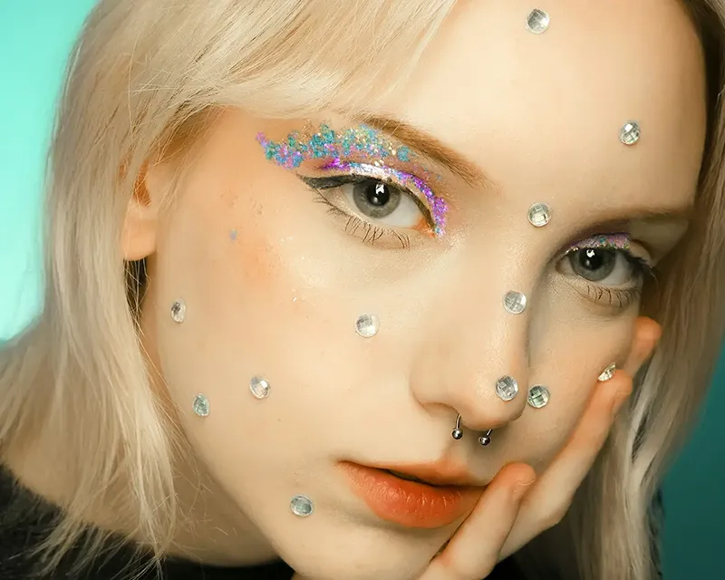 Lähikuva naisen kasvoista Studio Lumon kauneuspalveluissa, jossa kimaltava glitter-luomiväri ja suuret timanttikimalteet koristavat koko kasvojen ihoa.